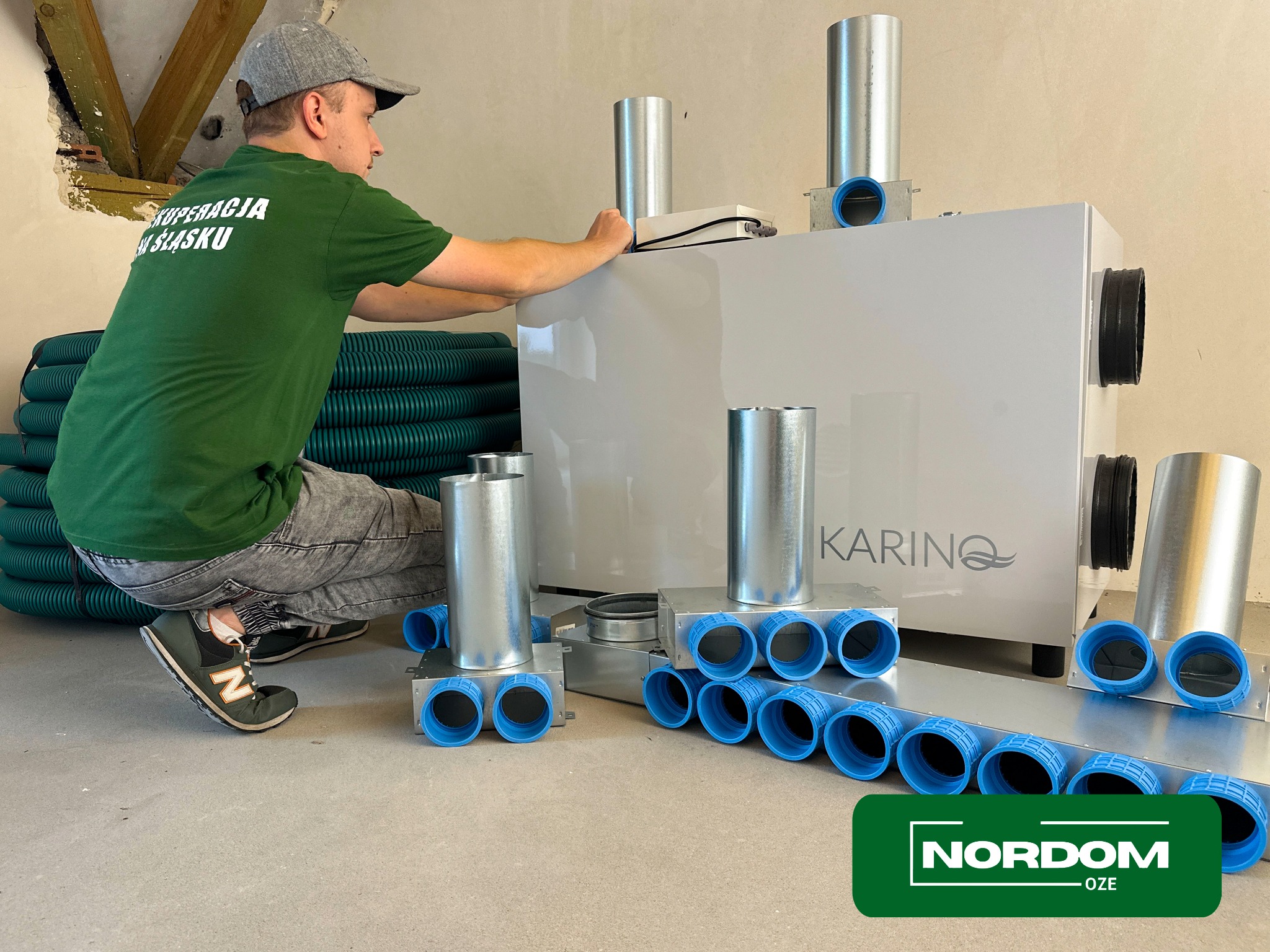 Montaż centrali wentylacyjnej z rekuperacją marki Karino przez instalatora w zielonej koszulce z napisem 'Rekuperacja na Śląsku'. Widoczne elementy instalacji, rury wentylacyjne w tle i logo Nordom...