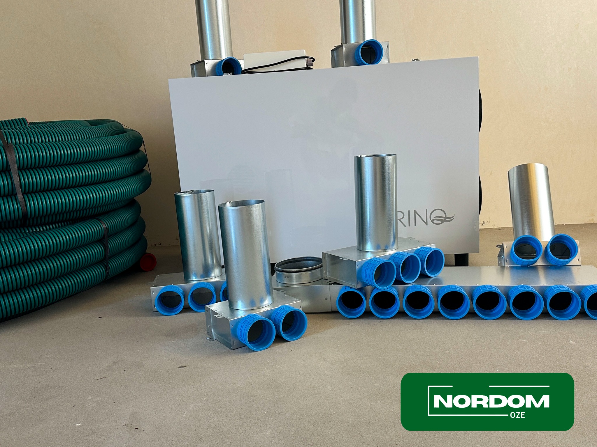 Centrala wentylacyjna z rekuperacją, rury wentylacyjne i kształtki na podłodze, widoczne logo firmy NORDOM OZE
