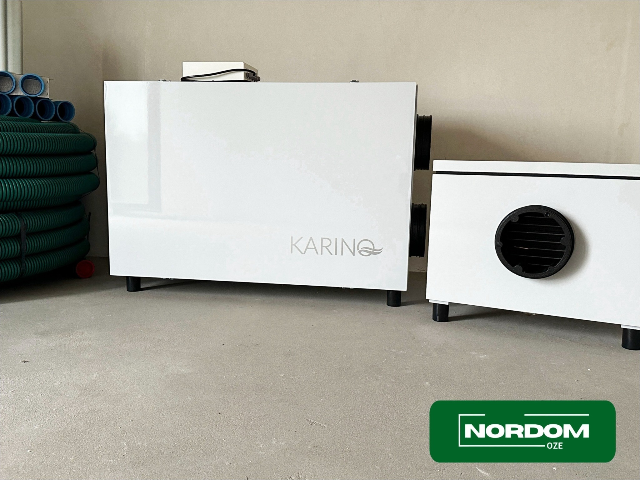 Białe urządzenia rekuperacyjne KARINO z czarnymi wlotami powietrza i stosem zielonych rur wentylacyjnych na surowym betonowym podłożu.