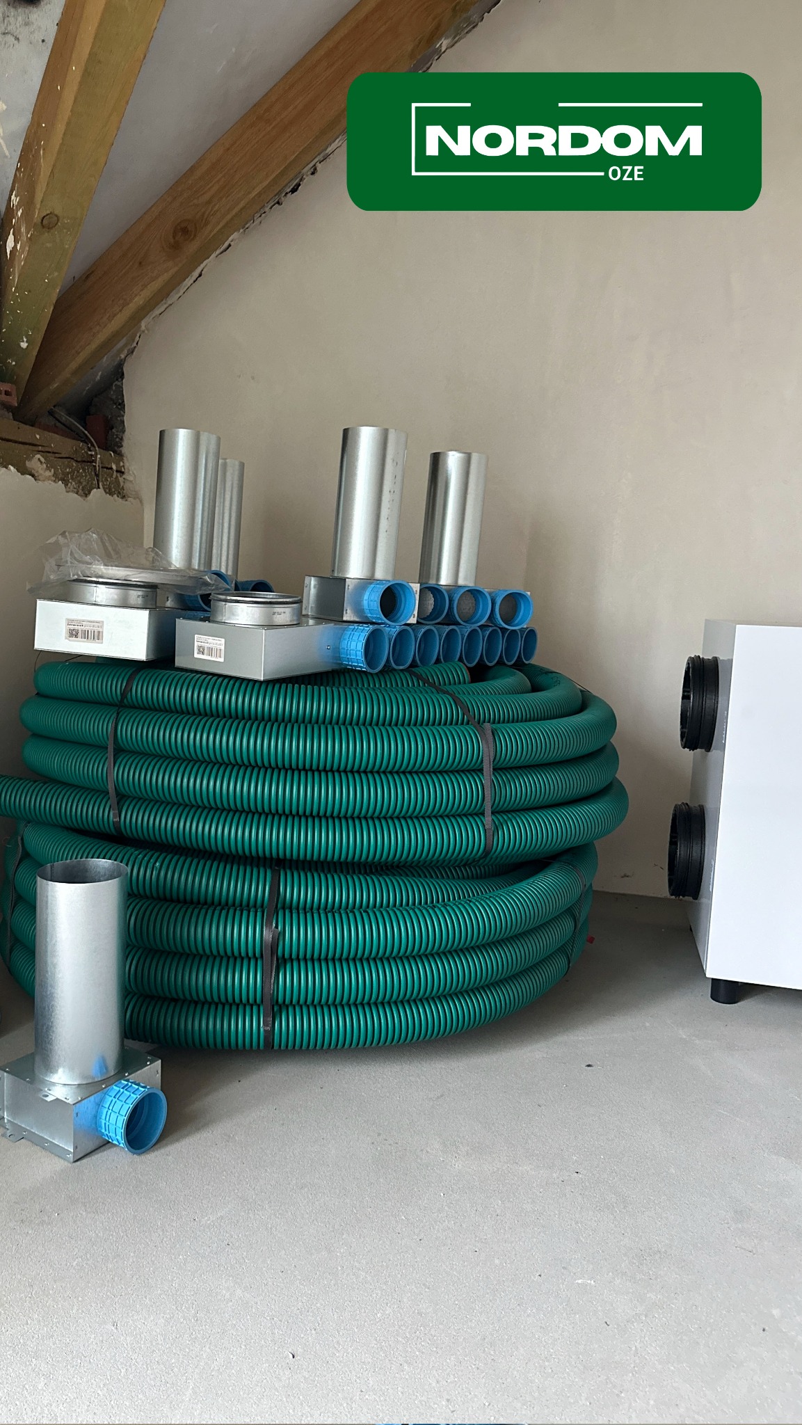 Elementy instalacji wentylacyjnej: zwój zielonych rur karbowanych, srebrne rury i kształtki, biała centrala wentylacyjna na surowym podłożu i tle ściany w trakcie wykończenia.