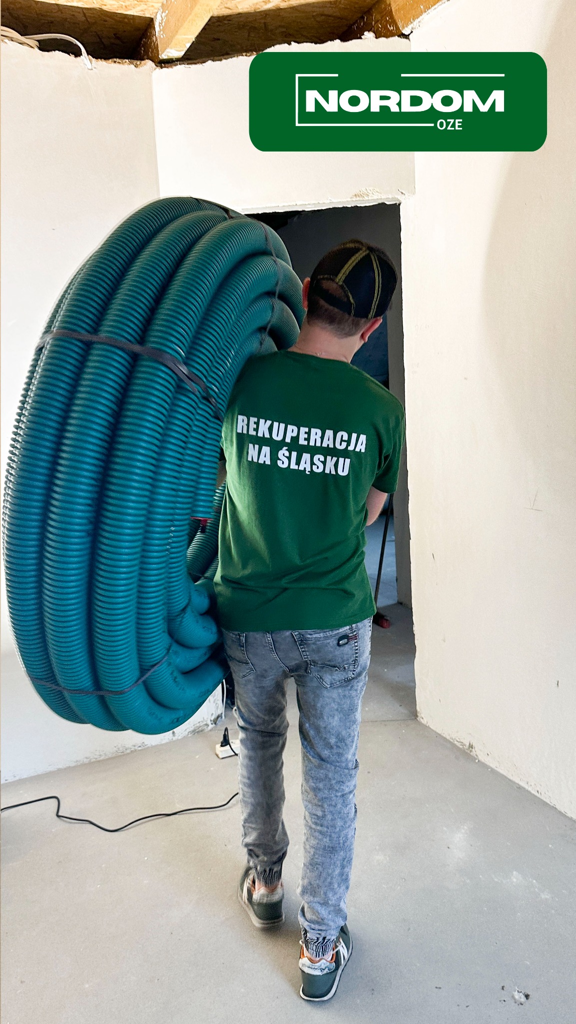 Mężczyzna w koszulce z napisem 'Rekuperacja na Śląsku' niesie na ramieniu dużą rolkę zielonych rur wentylacyjnych w surowym wnętrzu budynku.