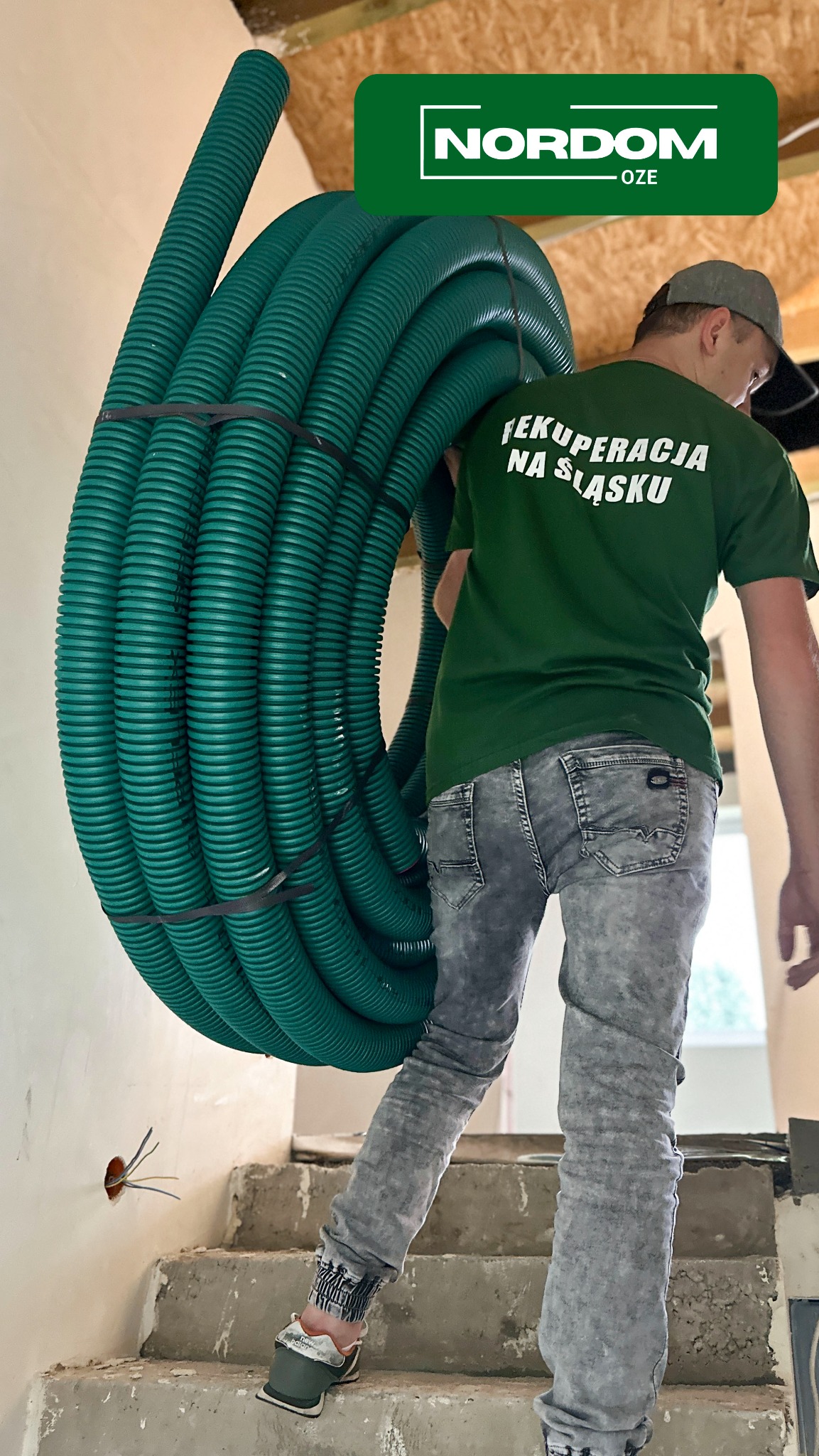 Instalator w zielonej koszulce z napisem 'Rekuperacja na Śląsku' niesie na ramieniu zwój zielonych rur do rekuperacji, w tle surowe schody i wystające kable elektryczne.