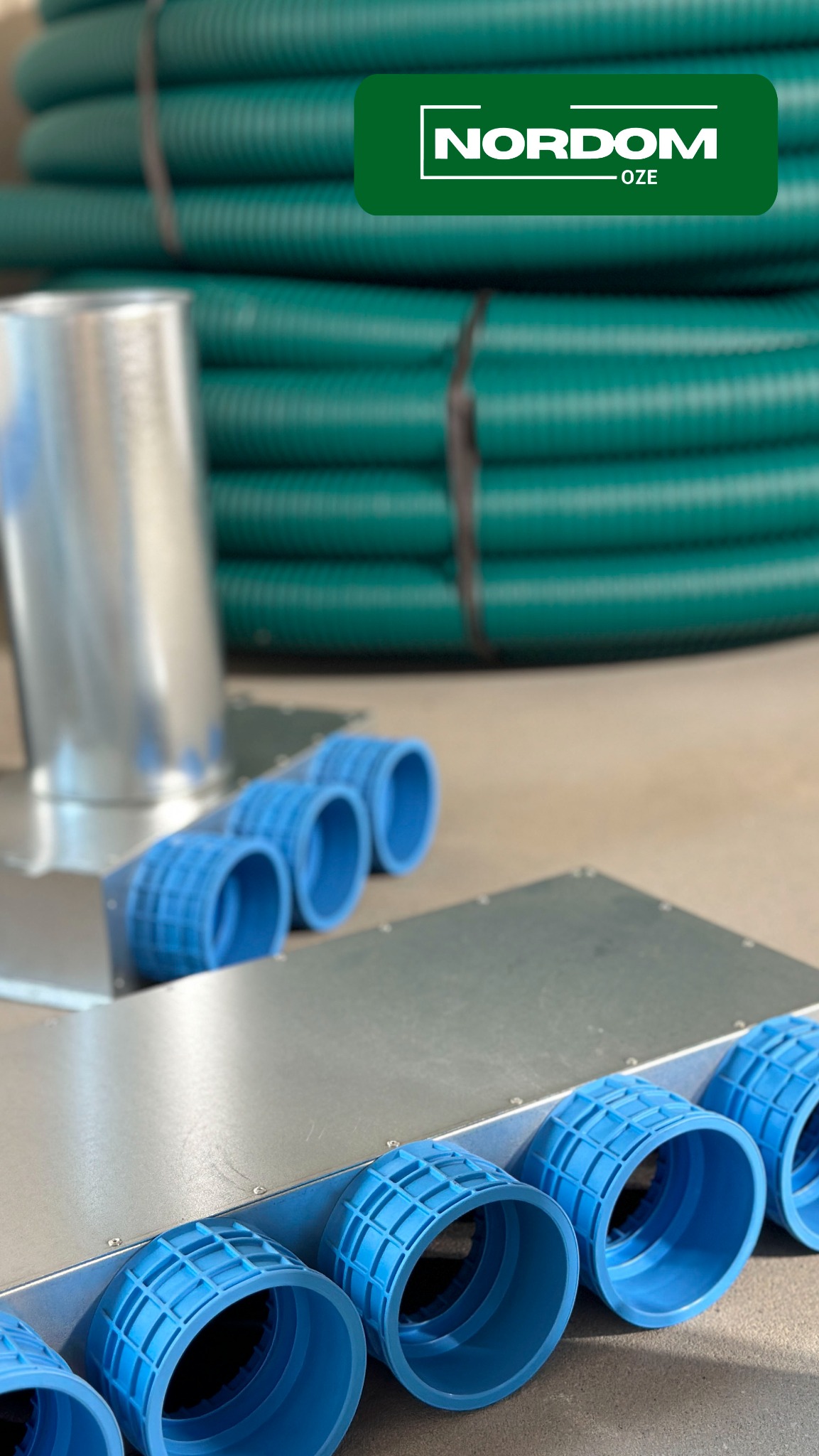 Elementy systemu wentylacyjnego: srebrna skrzynka z niebieskimi, okrągłymi wlotami powietrza z tworzywa sztucznego, w tle zwój zielonych rur wentylacyjnych i logo NORDOM-OZE.