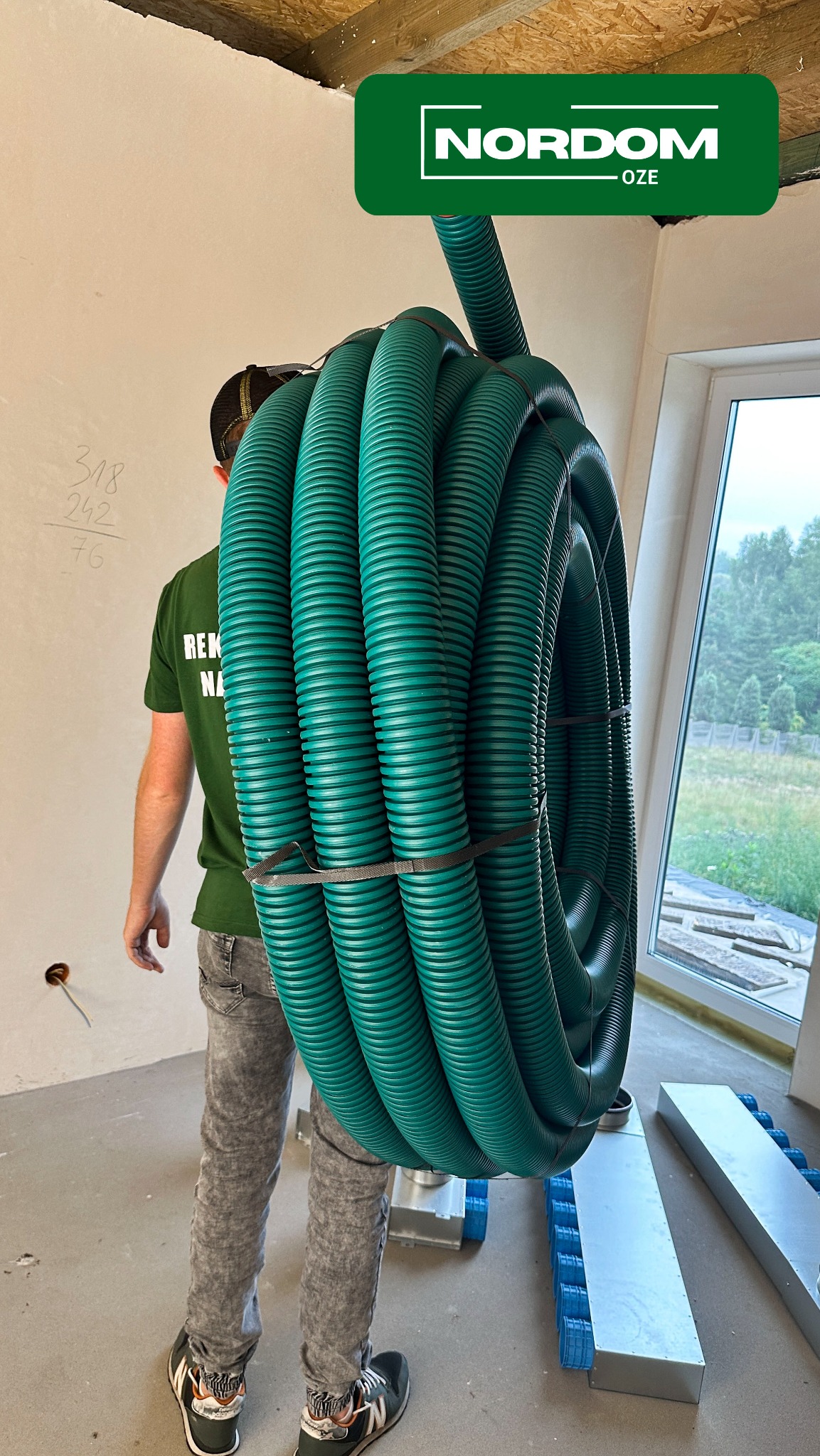Instalator niesie zwój zielonych, karbowanych rur wentylacyjnych na ramieniu w surowym wnętrzu budynku, obok widoczne kanały wentylacyjne i okno.