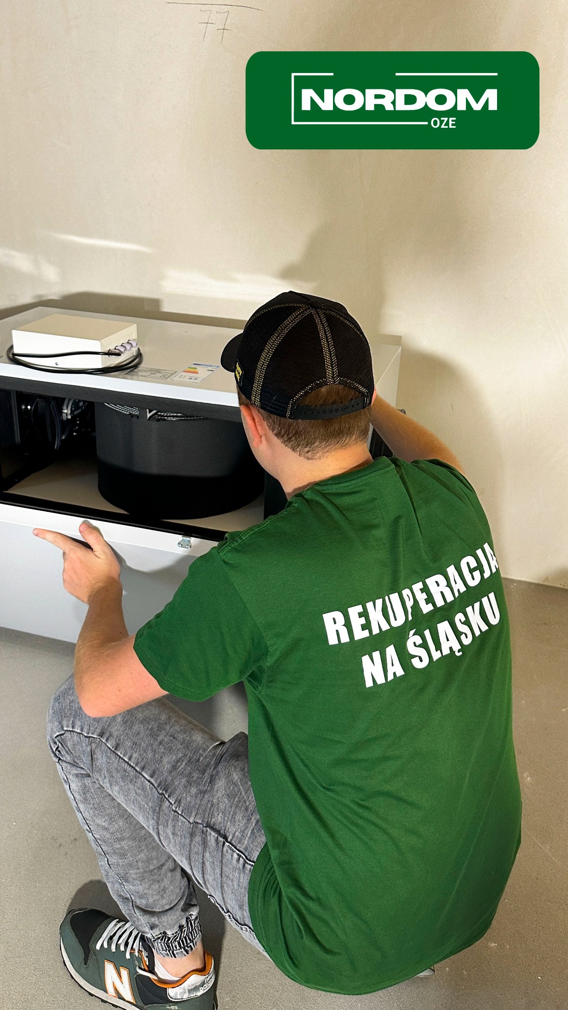 Instalator w zielonej koszulce z napisem 'Rekuperacja na Śląsku' kuca przed białą jednostką rekuperacyjną podczas montażu, widok z tyłu, na ścianie widoczny napis '77'.