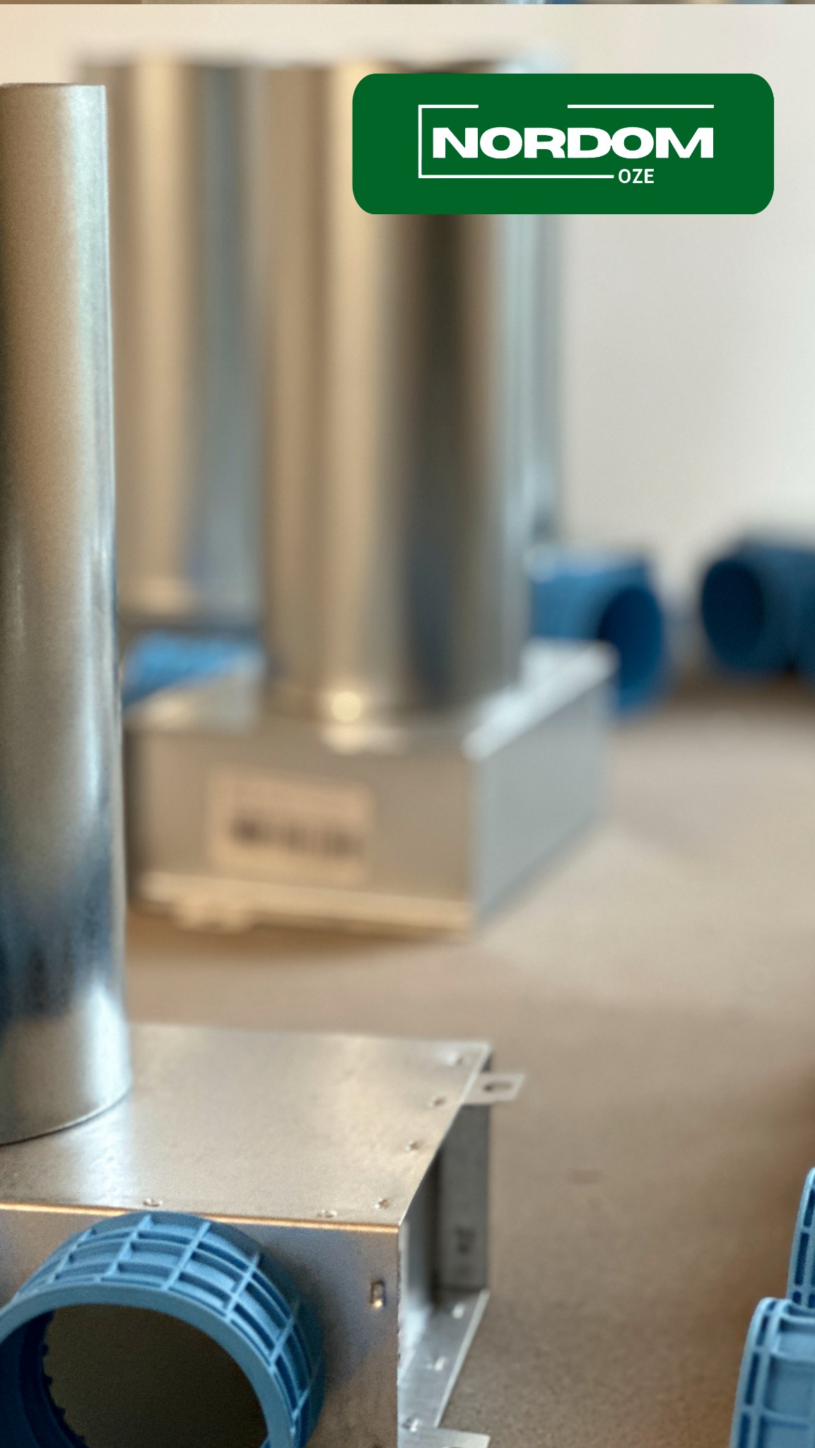 Elementy instalacji wentylacyjnej z blachy ocynkowanej i niebieskiego tworzywa sztucznego, widoczne rury i skrzynki, w tle logo firmy NORDOM-OZE.