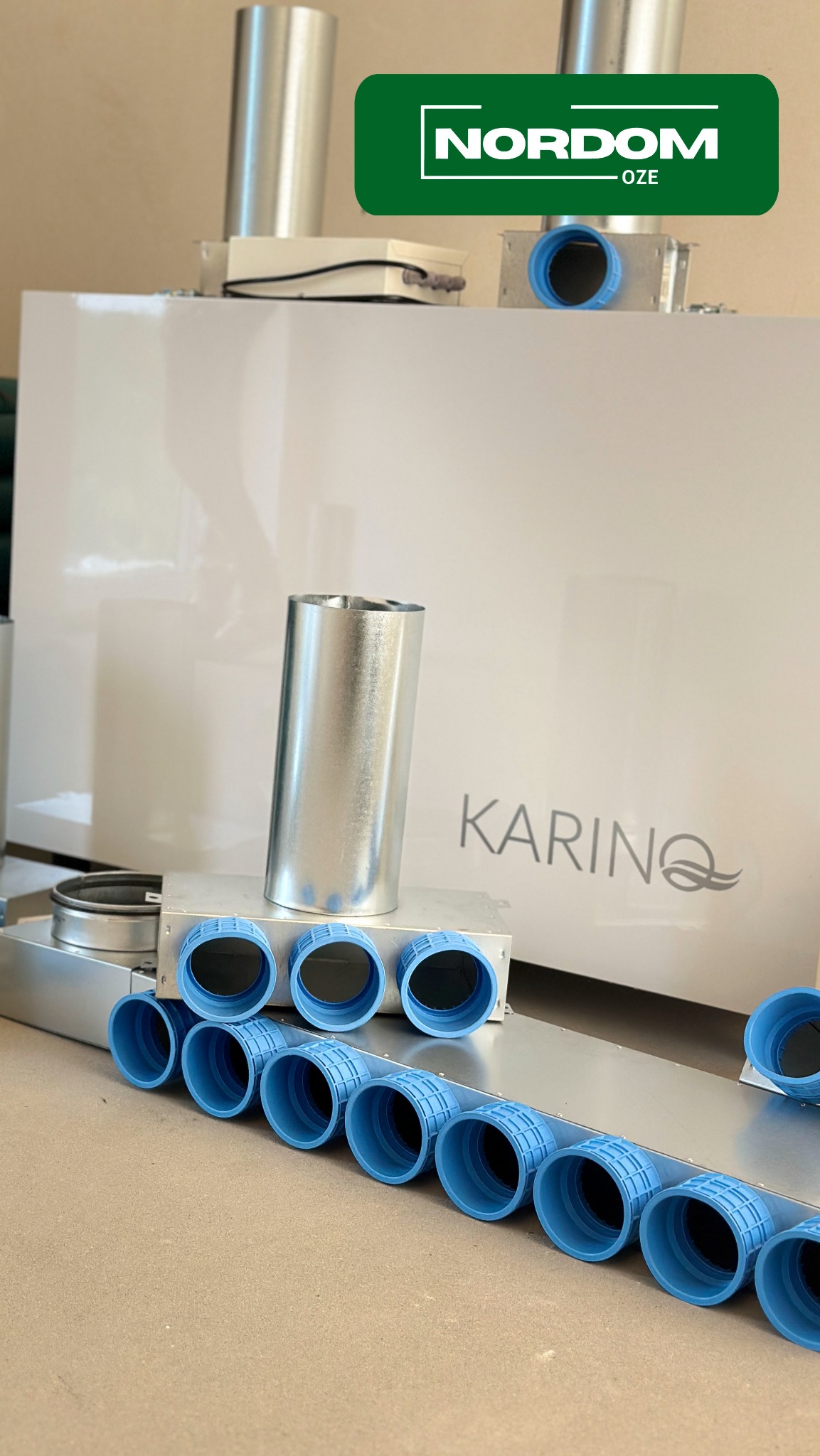 Biała centrala wentylacyjna z logo KARINQ, widoczne elementy instalacji: metalowe rury i niebieskie złączki, na górze logo NORDOM OZE.