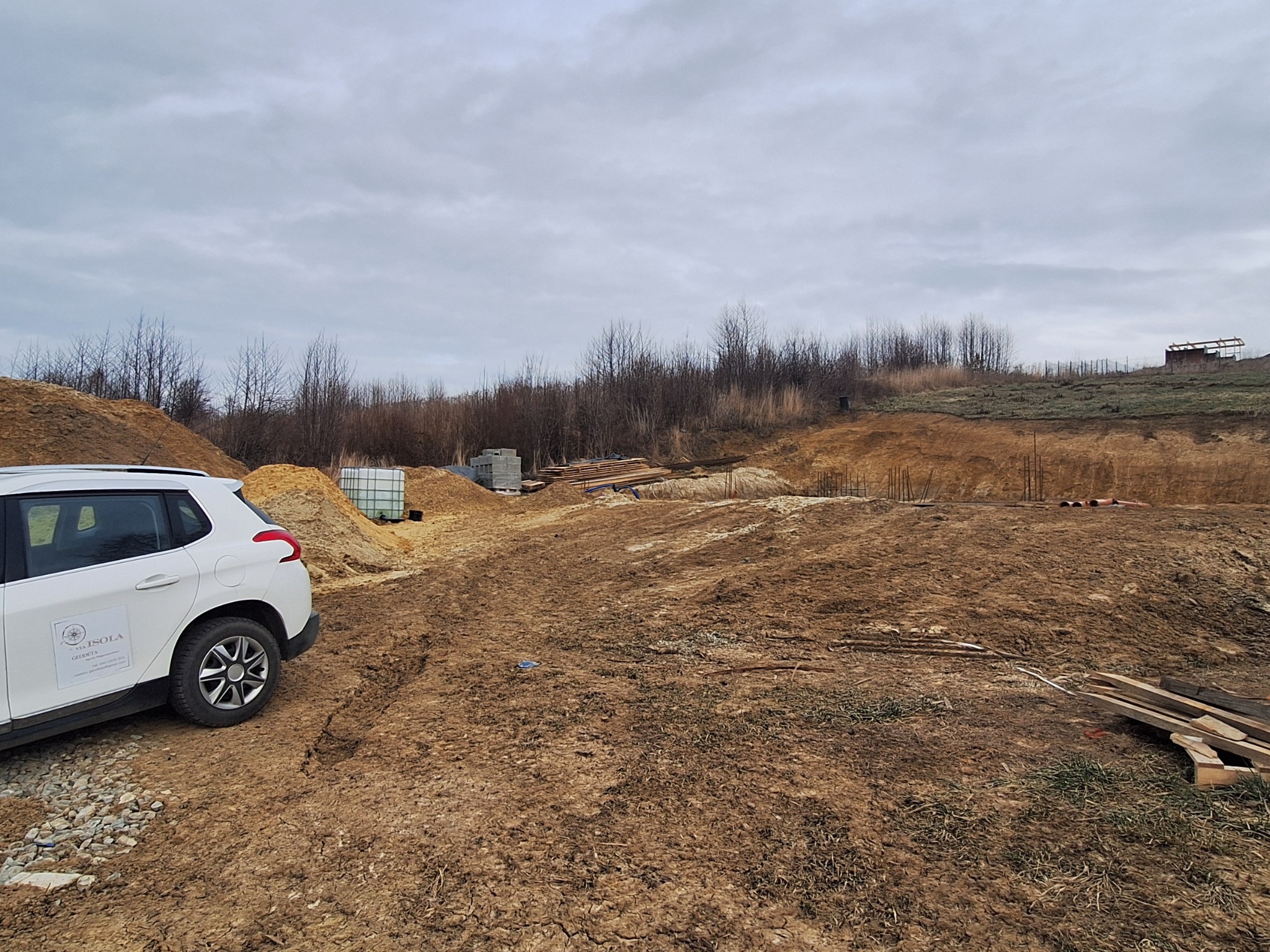 Biały samochód z logo firmy geodezyjnej na tle przygotowywanego terenu budowy, widoczne wykopane fundamenty i składowane materiały.