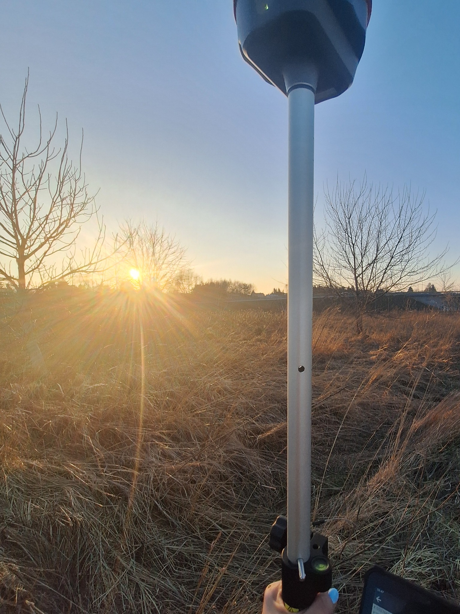 Pomiarowy odbiornik GNSS zamontowany na tyczce w terenie z widokiem na zachodzące słońce