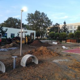 STORM Szymon Bujarek - Plac budowy z koparką i elementami placu zabaw w tle, oświetlony lampą uliczną. Widoczne betonowe tunele i przygotowane podłoże.