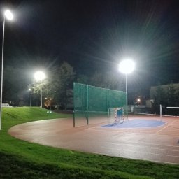 STORM Szymon Bujarek - Oświetlony nocą kompleks sportowy z boiskiem, bramkami i koszem do koszykówki. Widoczne lampy oświetleniowe i fragmenty zieleni. Ujęcie z dołu.