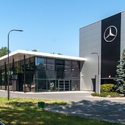 STORM Szymon Bujarek - Nowoczesny budynek salonu samochodowego Mercedes-Benz z przeszkloną fasadą i logo na elewacji, oświetlony lampami ulicznymi. Widok z zewnątrz, słoneczny dzień.