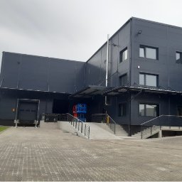 STORM Szymon Bujarek - Nowoczesny budynek przemysłowy z rampą i schodami w Przecieszynie. Widoczne elementy konstrukcyjne i oświetlenie zewnętrzne. Szara elewacja i kostka brukowa.