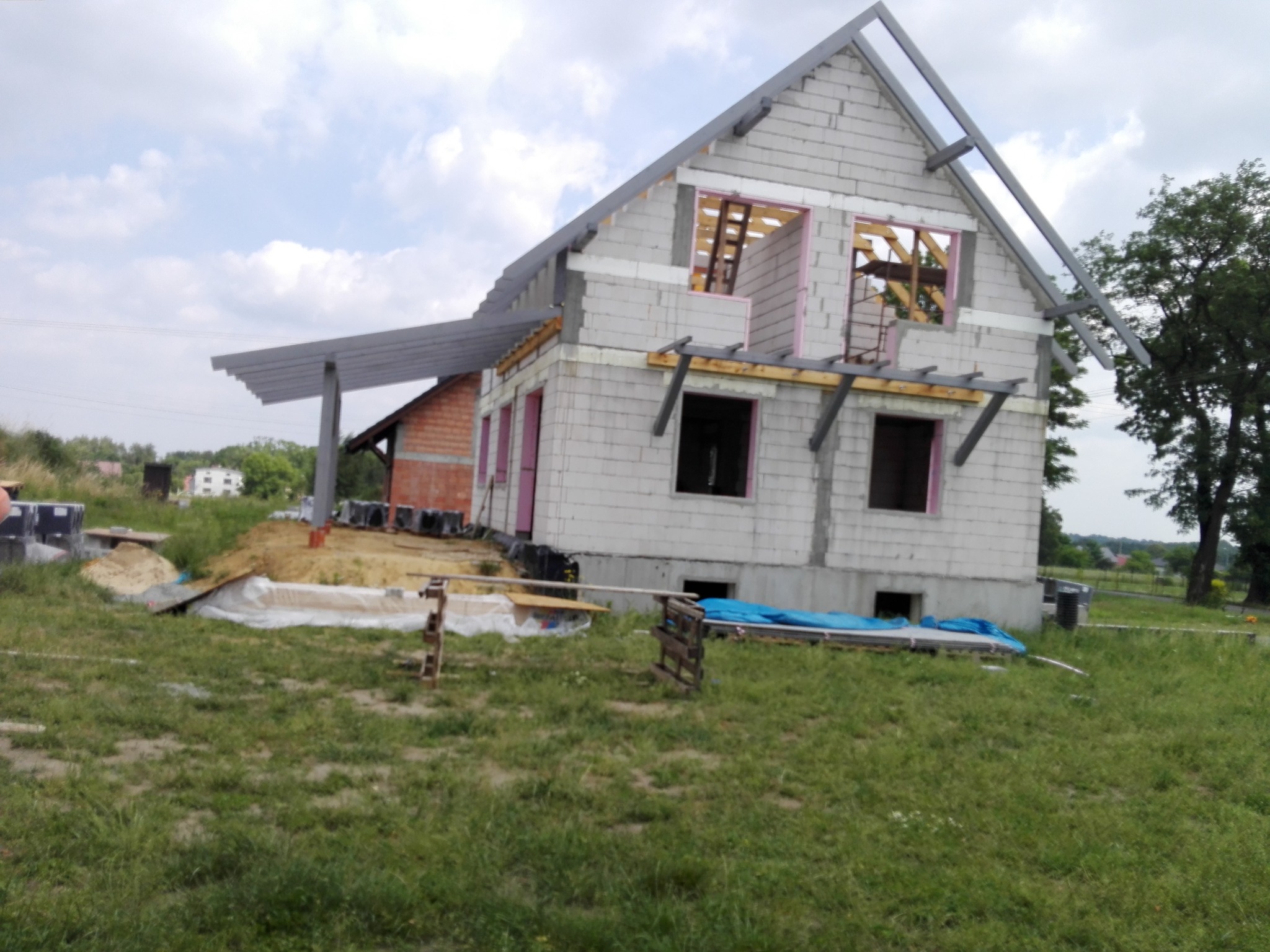 Surowy stan domu z betonu komórkowego w trakcie budowy, widoczne okna z różową izolacją, konstrukcja dachu i częściowo zadaszony taras. Otoczenie to trawa i drzewa.