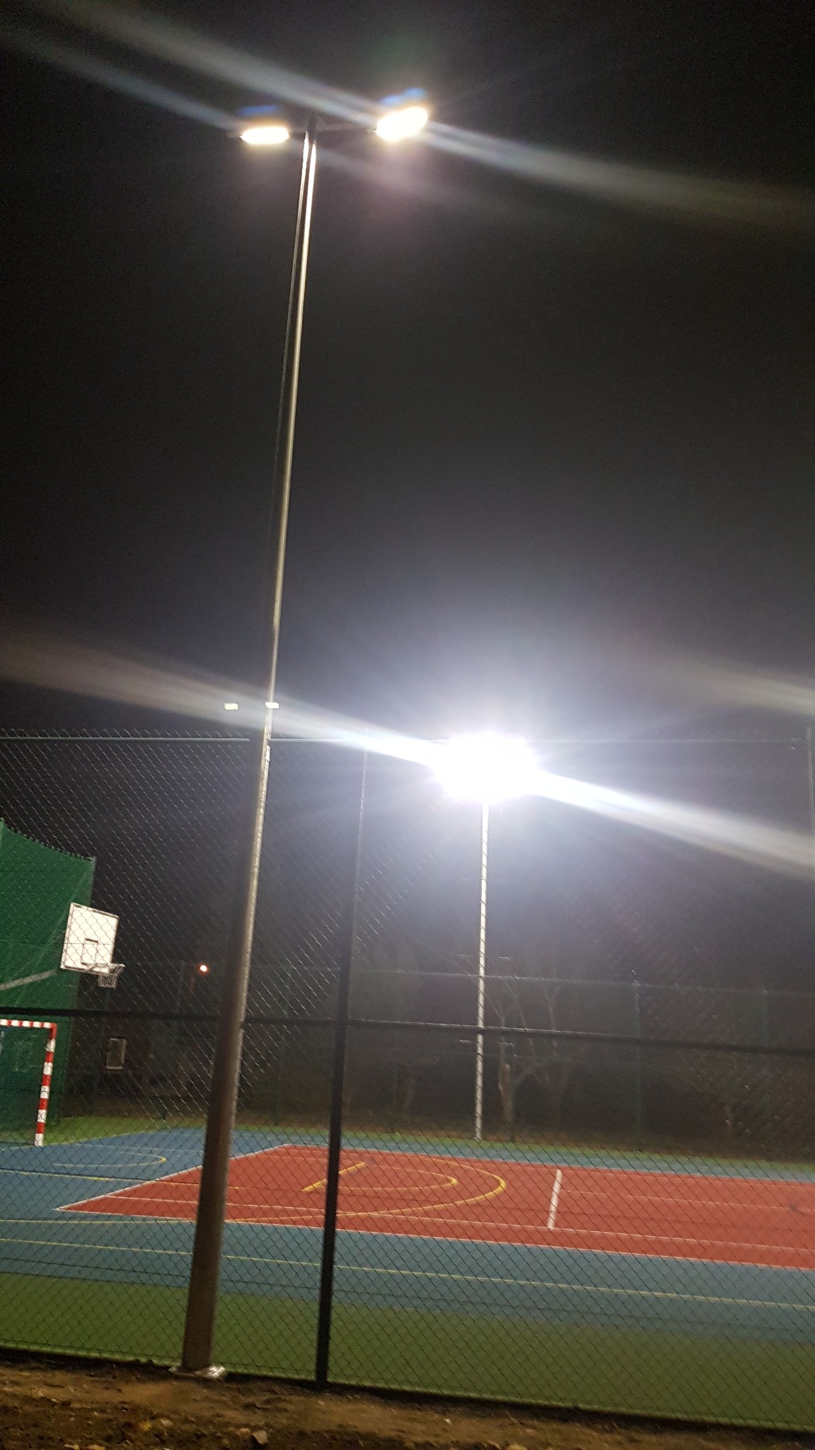 Oświetlony nocą, ogrodzony siatką, wielofunkcyjny plac sportowy z koszem i bramką. Widoczne dwie wysokie lampy oświetleniowe.