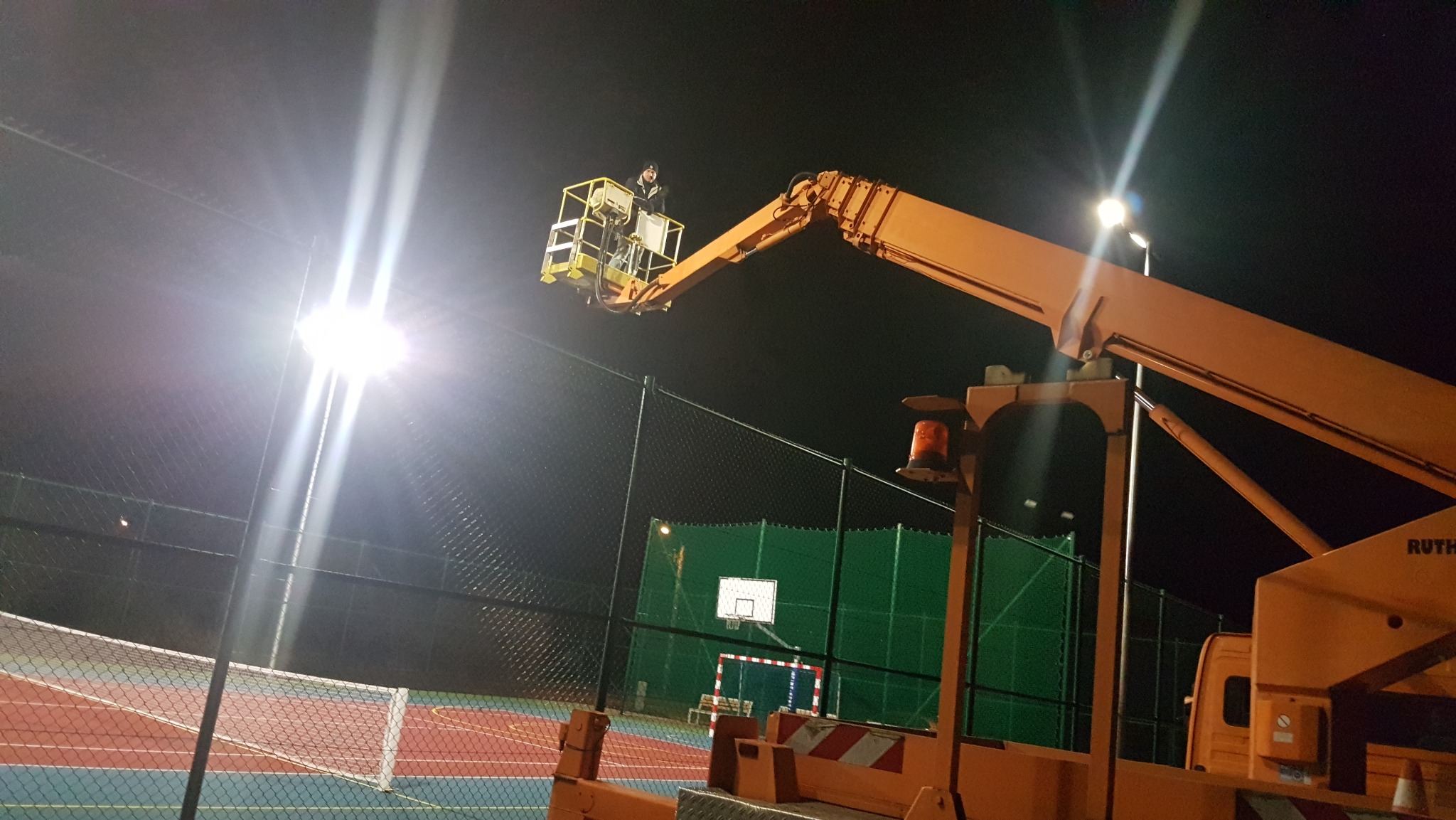 Pracownik na podnośniku koszowym montuje oświetlenie boiska sportowego nocą. Widoczne jaskrawe światła i siatka ogrodzeniowa.