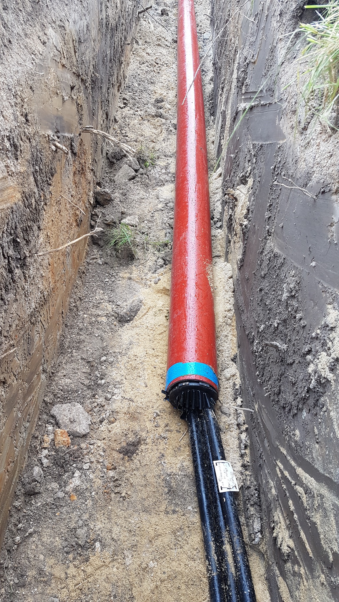 Czerwona rura osłonowa z niebieskim oznaczeniem, częściowo zakopana w wykopie ziemnym, chroniąca czarne kable elektryczne. Widok z góry na instalację.