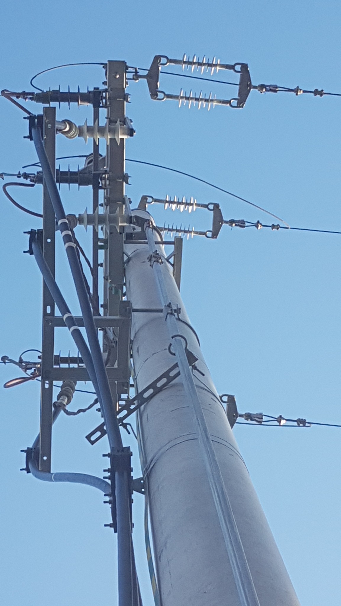 Słup energetyczny z przewodami i izolatorami, widok z dołu na tle błękitnego nieba. Elementy konstrukcyjne i kable w zbliżeniu.