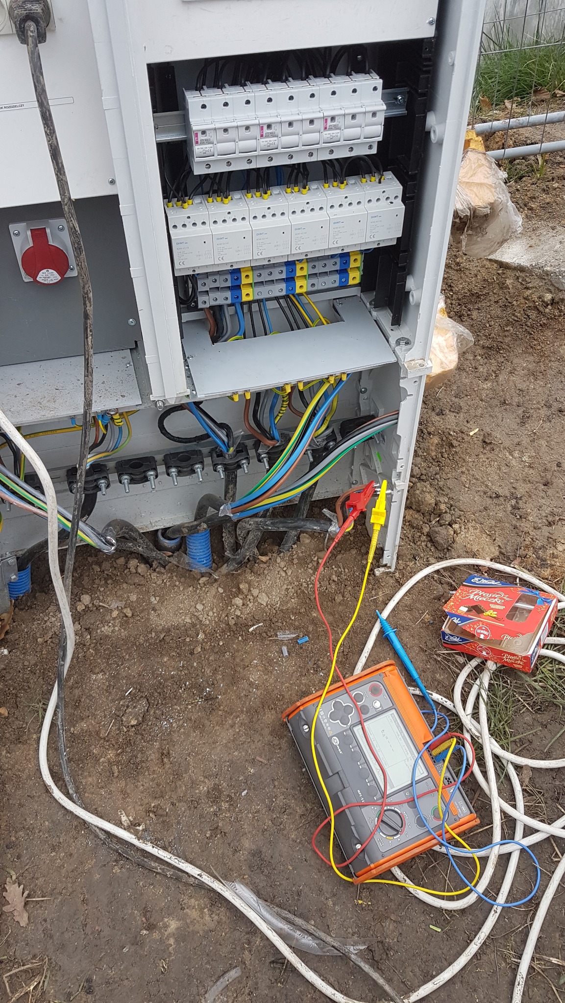 Otwarta skrzynka elektryczna z widocznymi przewodami, bezpiecznikami i miernikiem elektrycznym na ziemi. Widoczna skrzynka czekoladek.