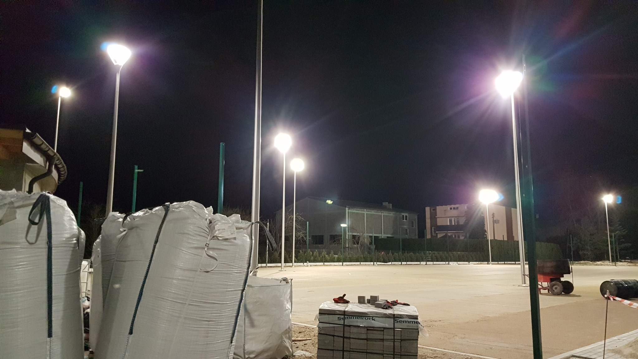 Nocny widok na plac z nowo zamontowanym oświetleniem. Widoczne lampy uliczne, worki z materiałami budowlanymi i budynek w tle. Oświetlenie placu po zmroku.