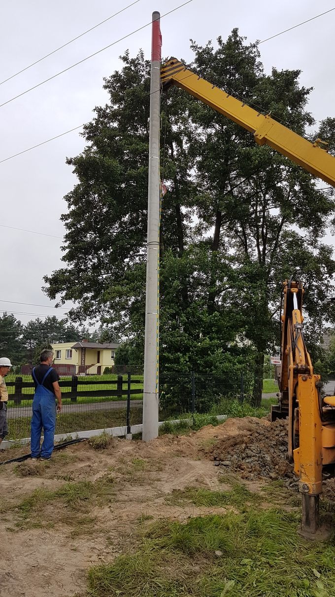 Montaż słupa oświetleniowego z użyciem dźwigu. Widoczni pracownicy w strojach roboczych i koparka. Słup zabezpieczony czerwoną folią na górze. Prace ziemne w toku.