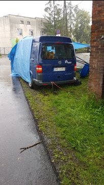 Niebieski bus VW Transporter, częściowo przykryty plandeką, zaparkowany na trawniku obok chodnika i budynku. Widoczne tablice rejestracyjne i logo firmy.