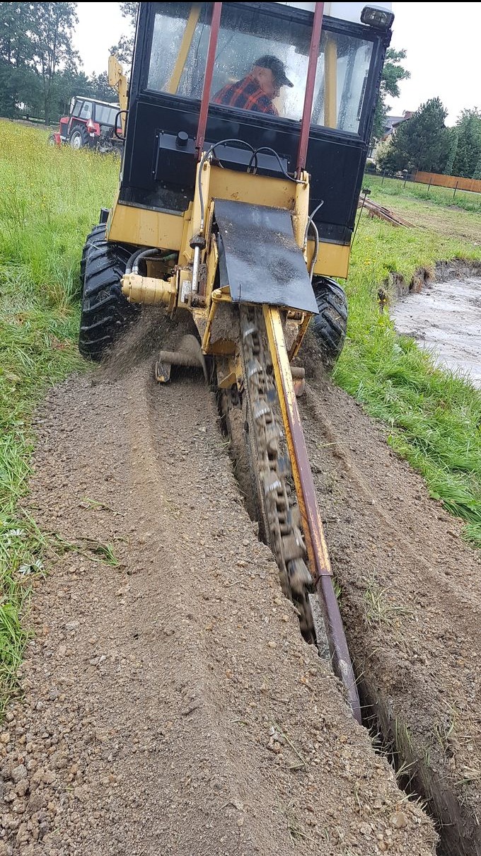 Żółta koparka łańcuchowa w trakcie pracy, wykopuje rów w ziemi w Przecieszynie. W tle widoczny traktor i zabudowania.