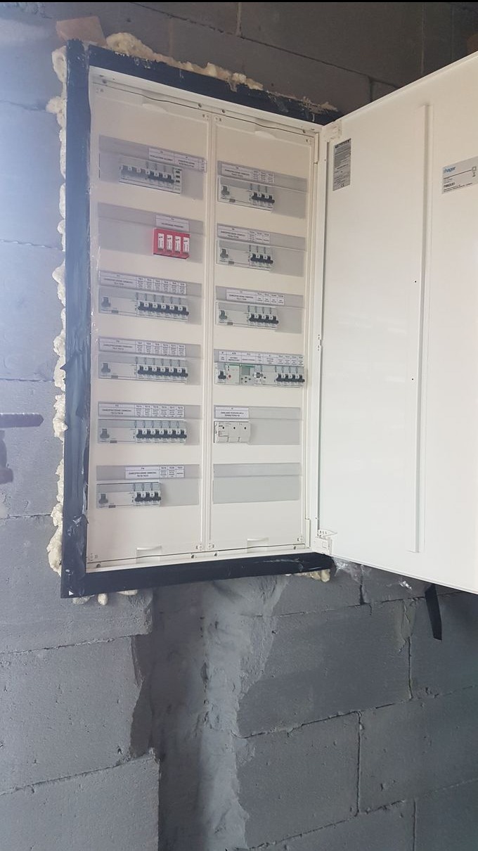 Otwarta, biała skrzynka rozdzielcza z bezpiecznikami, zamontowana na szarej, betonowej ścianie. Widoczne czarne obramowanie i pianka montażowa.