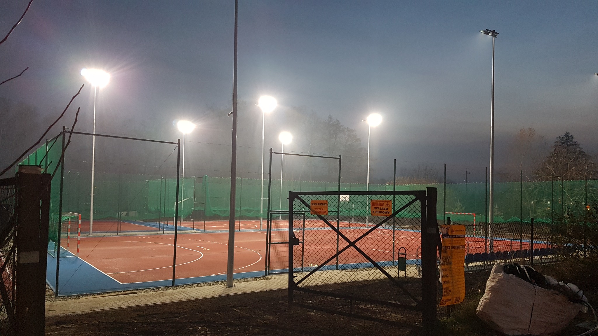 Oświetlony, wielofunkcyjny plac sportowy z bramkami i siatką, widoczny przez otwartą bramę. Wysokie lampy oświetlają boisko w wieczornej scenerii.