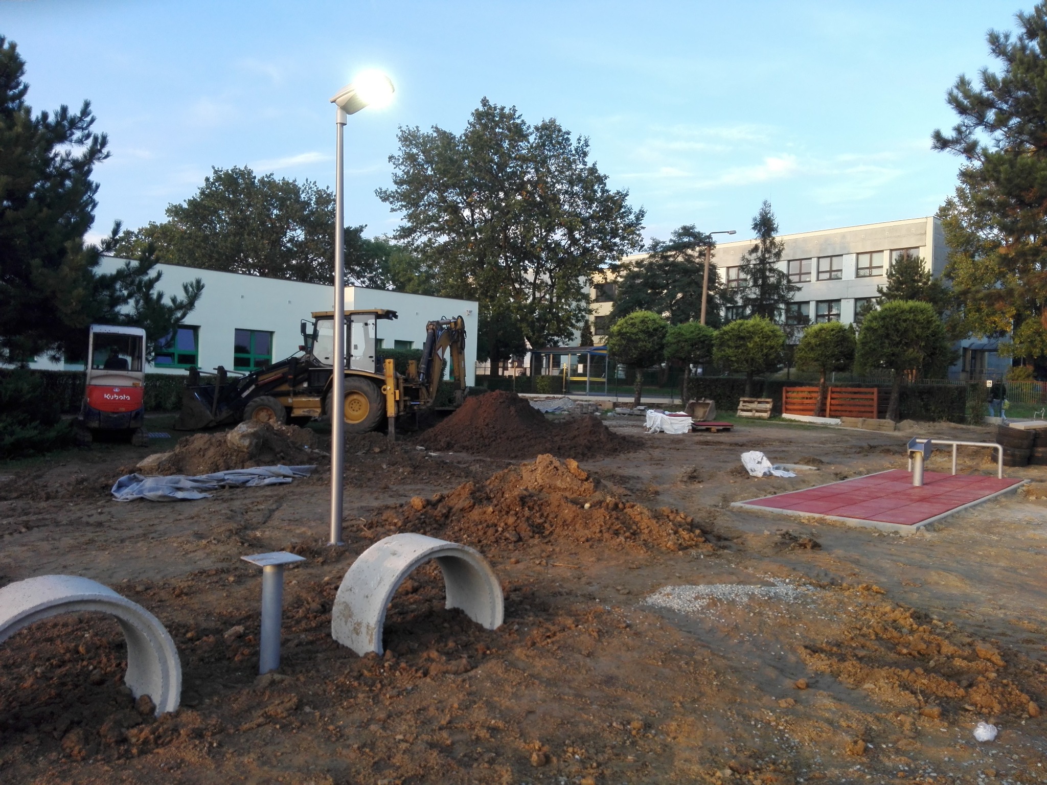 Plac budowy z koparką i elementami placu zabaw w tle, oświetlony lampą uliczną. Widoczne betonowe tunele i przygotowane podłoże.