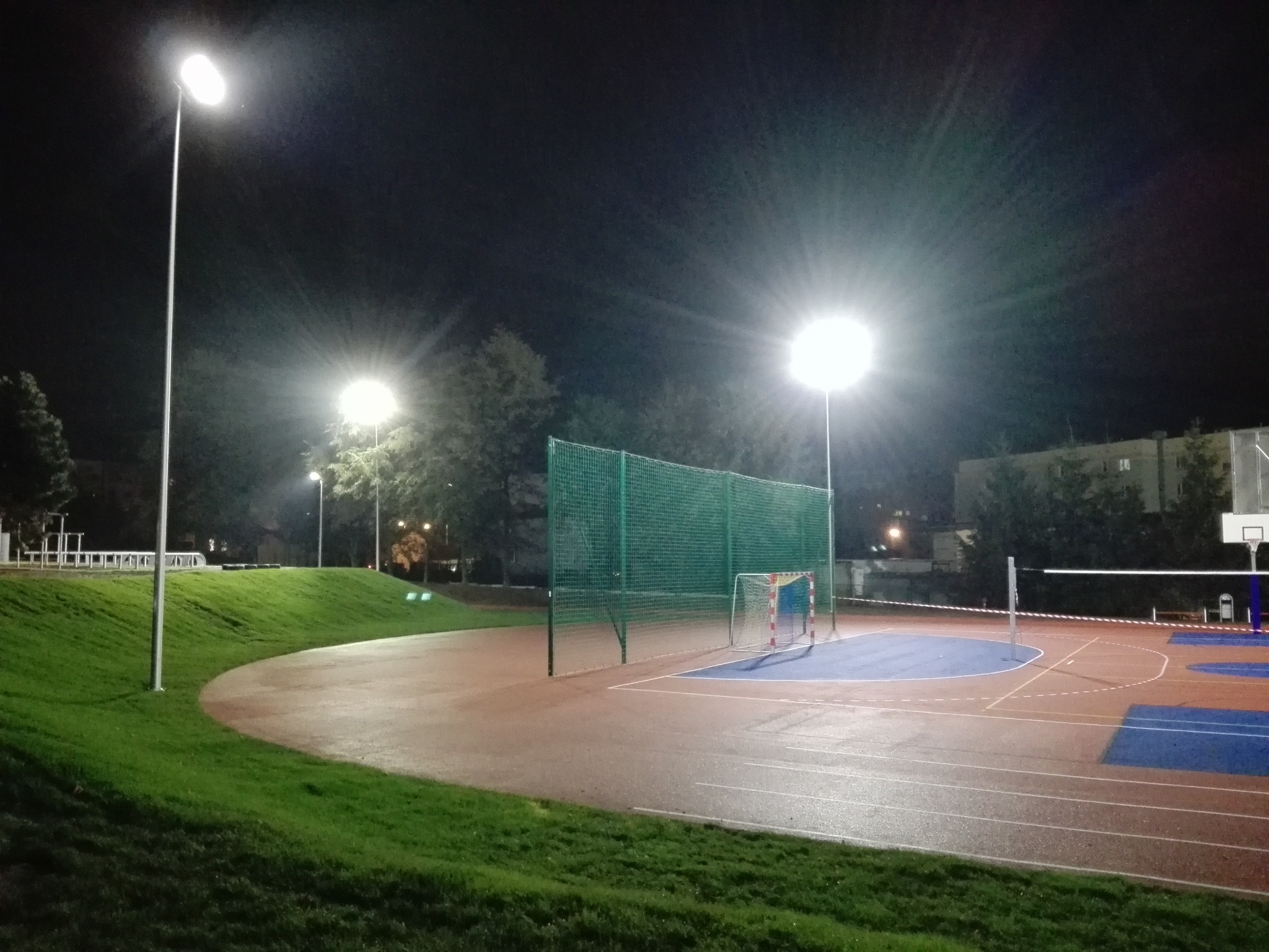 Oświetlony nocą kompleks sportowy z boiskiem, bramkami i koszem do koszykówki. Widoczne lampy oświetleniowe i fragmenty zieleni. Ujęcie z dołu.
