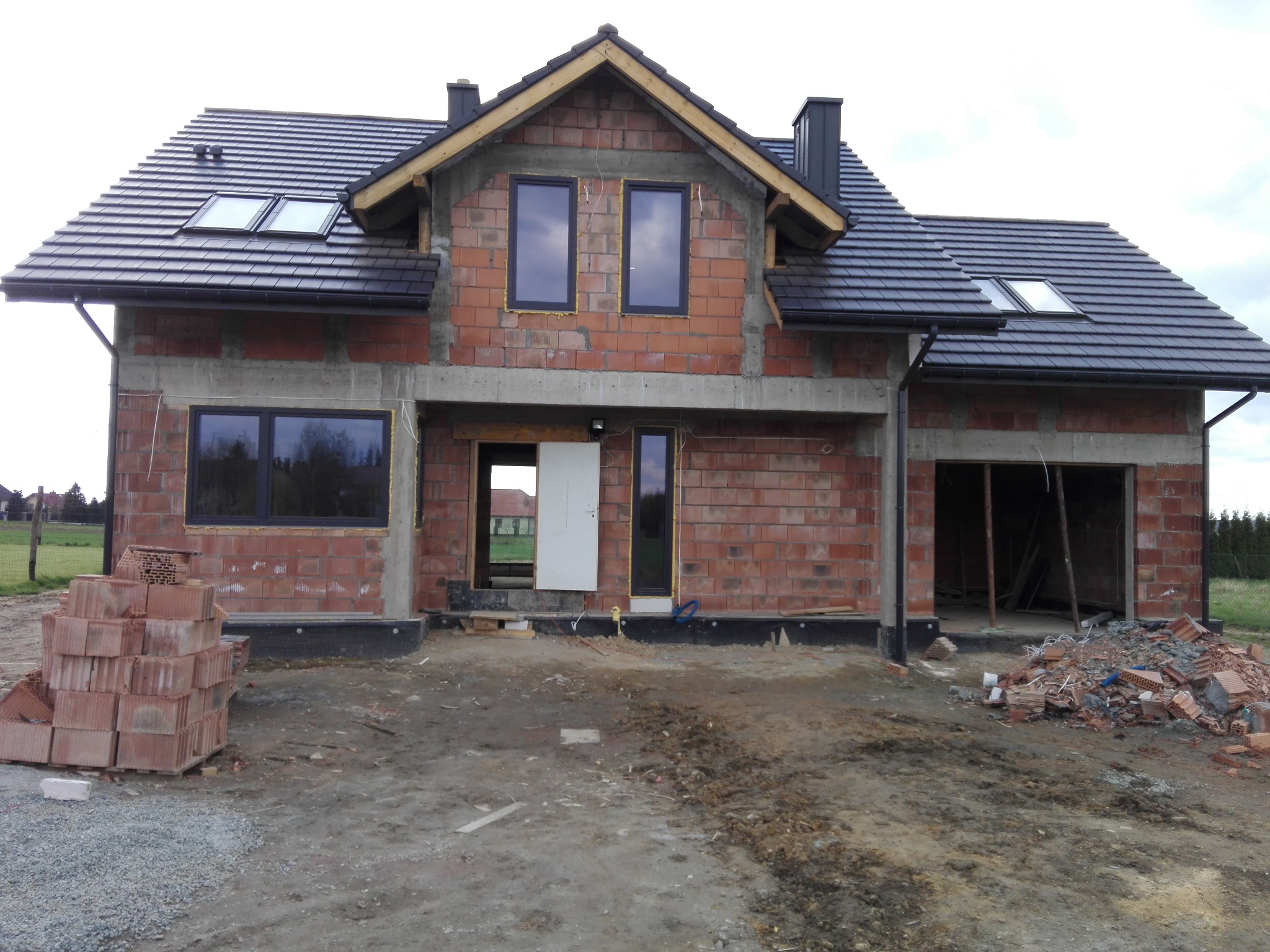 Surowy stan domu jednorodzinnego z czerwonej cegły z czarną dachówką, widoczne okna i drzwi, teren budowy z gruzem i stertą cegieł. Etap budowy.