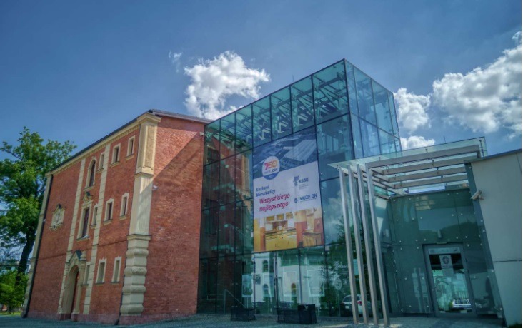 Elewacja budynku z czerwonej cegły połączona z nowoczesną, szklaną konstrukcją. Widoczne okna, błękitne niebo z chmurami i fragment zieleni. Ujęcie z dołu.