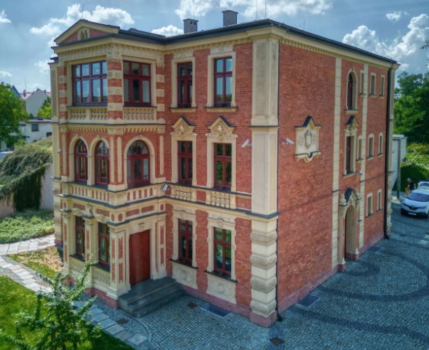 Elewacja odrestaurowanego, dwupiętrowego budynku z czerwonej cegły z kremowymi detalami architektonicznymi i ciemnoczerwoną stolarką okienną.