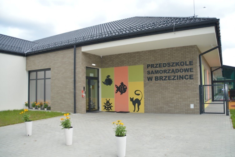 Nowoczesny budynek przedszkola z cegły klinkierowej, z kolorową dekoracją ścienną przedstawiającą zwierzęta. Widoczne kwiaty w donicach i fragment dachu.