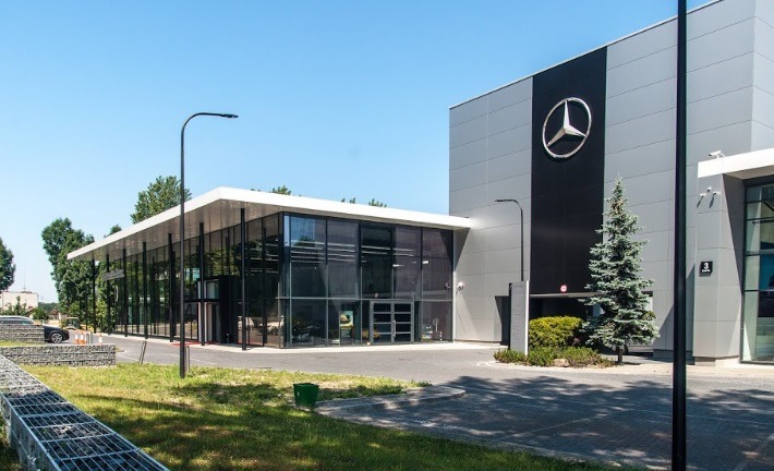 Nowoczesny budynek salonu samochodowego Mercedes-Benz z przeszkloną fasadą i logo na elewacji, oświetlony lampami ulicznymi. Widok z zewnątrz, słoneczny dzień.