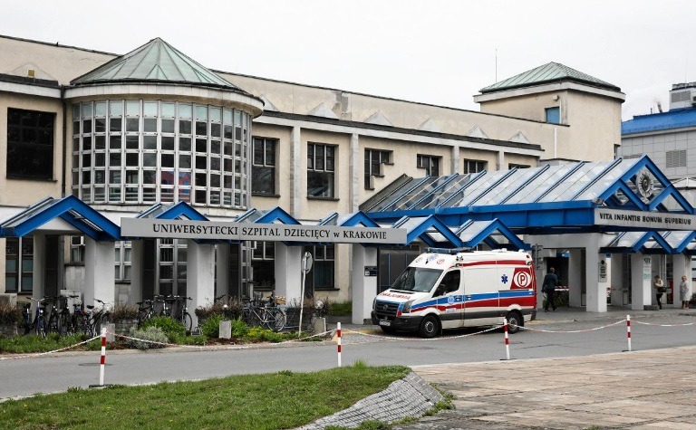 Szpital Uniwersytecki Dziecięcy w Krakowie z zaparkowaną karetką pogotowia. Widoczny budynek z przeszkloną kopułą i niebieskim zadaszeniem nad wejściem.
