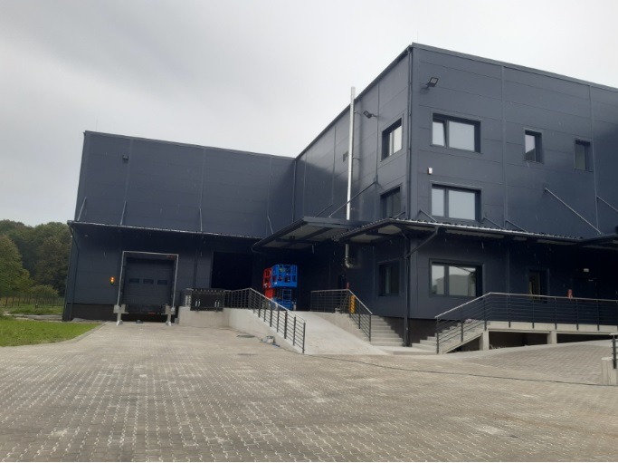 Nowoczesny budynek przemysłowy z rampą i schodami w Przecieszynie. Widoczne elementy konstrukcyjne i oświetlenie zewnętrzne. Szara elewacja i kostka brukowa.