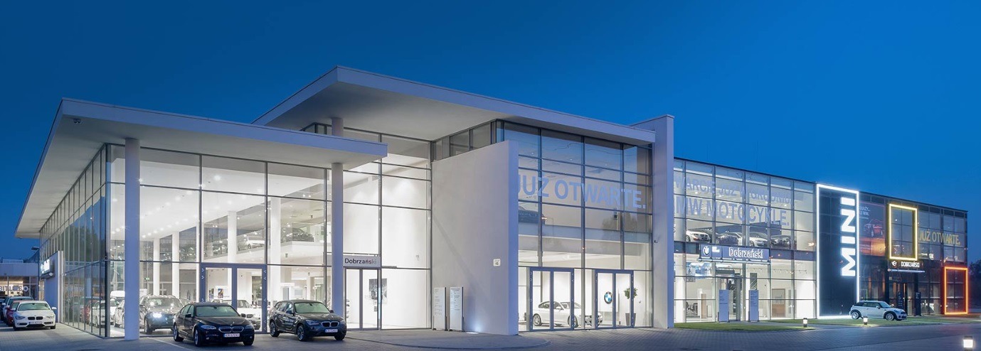 Nowoczesny salon samochodowy BMW i MINI z przeszkloną fasadą, oświetlony wieczorem. Widoczne samochody na zewnątrz i wewnątrz budynku. Minimalistyczna architektura.