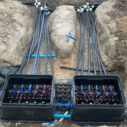 GreenEM Michał Flekiej - Dwa czarne pojemniki z zaworami i przewodami w wykopie ziemnym, niebieski klucz leży na ziemi obok przewodów, widoczne narzędzia na chodniku.