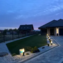 Oświetlona posesja z równo przystrzyżonym trawnikiem, ozdobnym kamieniem i ścieżką z kostki brukowej w odcieniach szarości. W tle widoczny dom i wieczorne niebo.