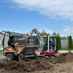 GreenEM Michał Flekiej - Żółta wywrotka i koparka na placu budowy. Operator koparki nabiera ziemię do wywrotki. W tle widoczne panele słoneczne i budynki mieszkalne.