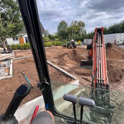 GreenEM Michał Flekiej - Widok z kabiny koparki na plac budowy: wykop pod fundamenty, przygotowany teren, sprzęt budowlany. Perspektywa operatora maszyny.