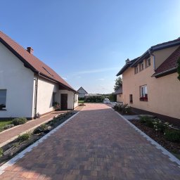 GreenEM Michał Flekiej - Ułożona kostka brukowa tworzy ścieżkę między dwoma domami. Widoczne rabaty z roślinami i trawnik. Słoneczny dzień, błękitne niebo. Kostka w odcieniach brązu i czerwieni.