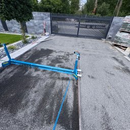 GreenEM Michał Flekiej - Niebieska profilarka do precyzyjnego rozprowadzania kruszywa na podjeździe, przygotowująca podłoże pod kostkę brukową, z nowoczesnym ogrodzeniem i zielenią w tle.