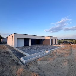 GreenEM Michał Flekiej - Nowoczesny dom z płaskim dachem i ciemnymi bramami garażowymi, przed którym ułożono ciemną kostkę brukową z betonowymi obrzeżami. Otoczenie to piaszczysty teren z widocznymi śladami opon...