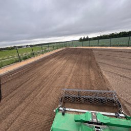 GreenEM Michał Flekiej - Rozległy teren z precyzyjnie wyrównaną i spulchnioną glebą, gotową pod nasadzenia, z zielonym agregatem uprawowym w prawym dolnym rogu. W tle siatkowe ogrodzenie.