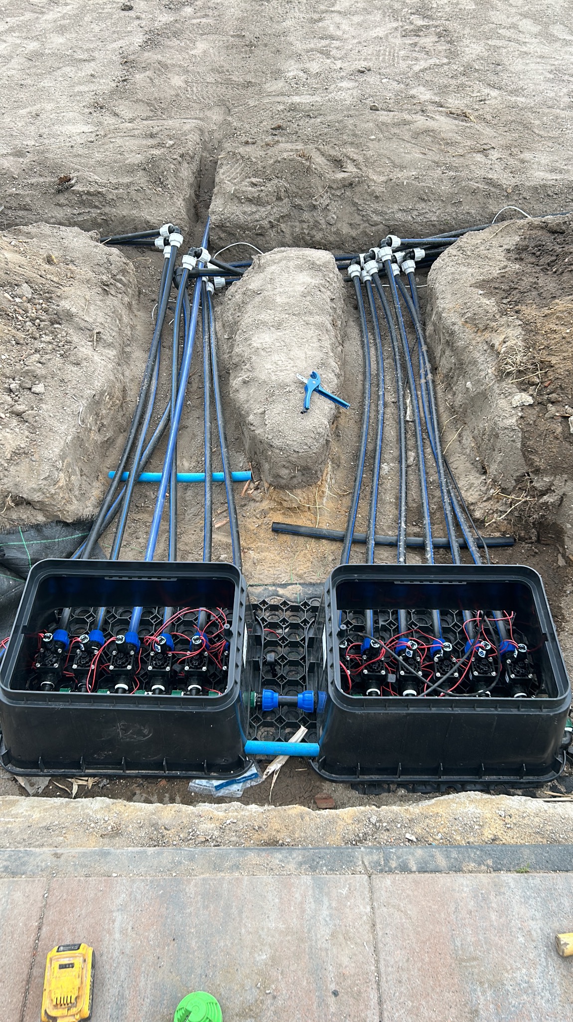 Dwa czarne pojemniki z zaworami i przewodami w wykopie ziemnym, niebieski klucz leży na ziemi obok przewodów, widoczne narzędzia na chodniku.