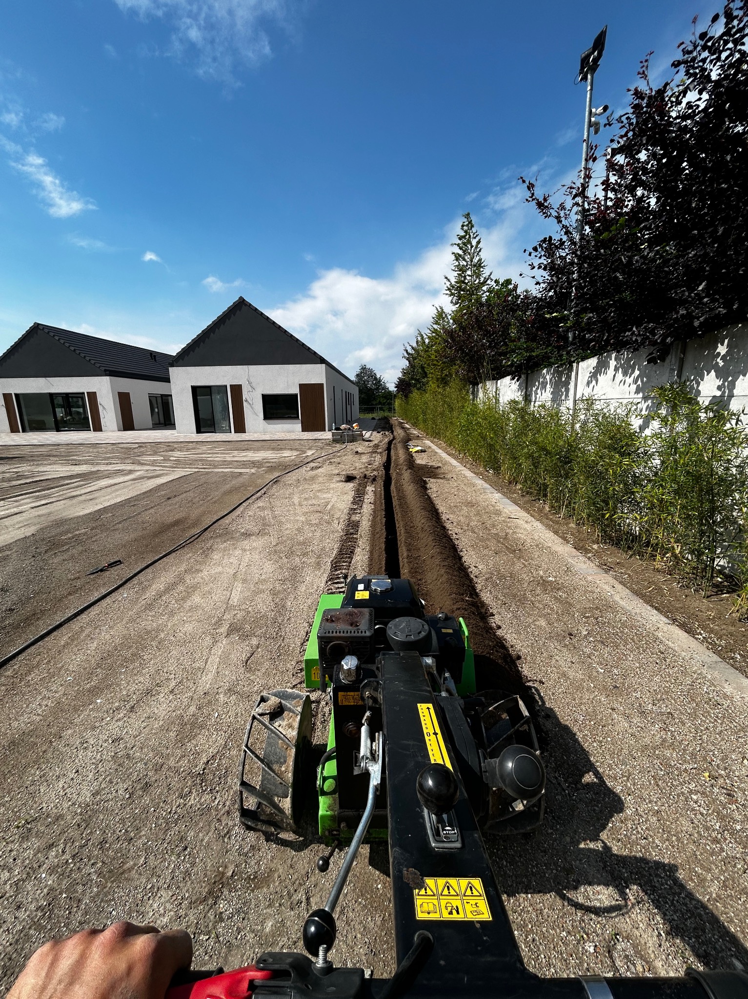 Wykonywanie wykopu pod system nawadniania za pomocą glebogryzarki, widoczne dwa nowoczesne domy w tle.