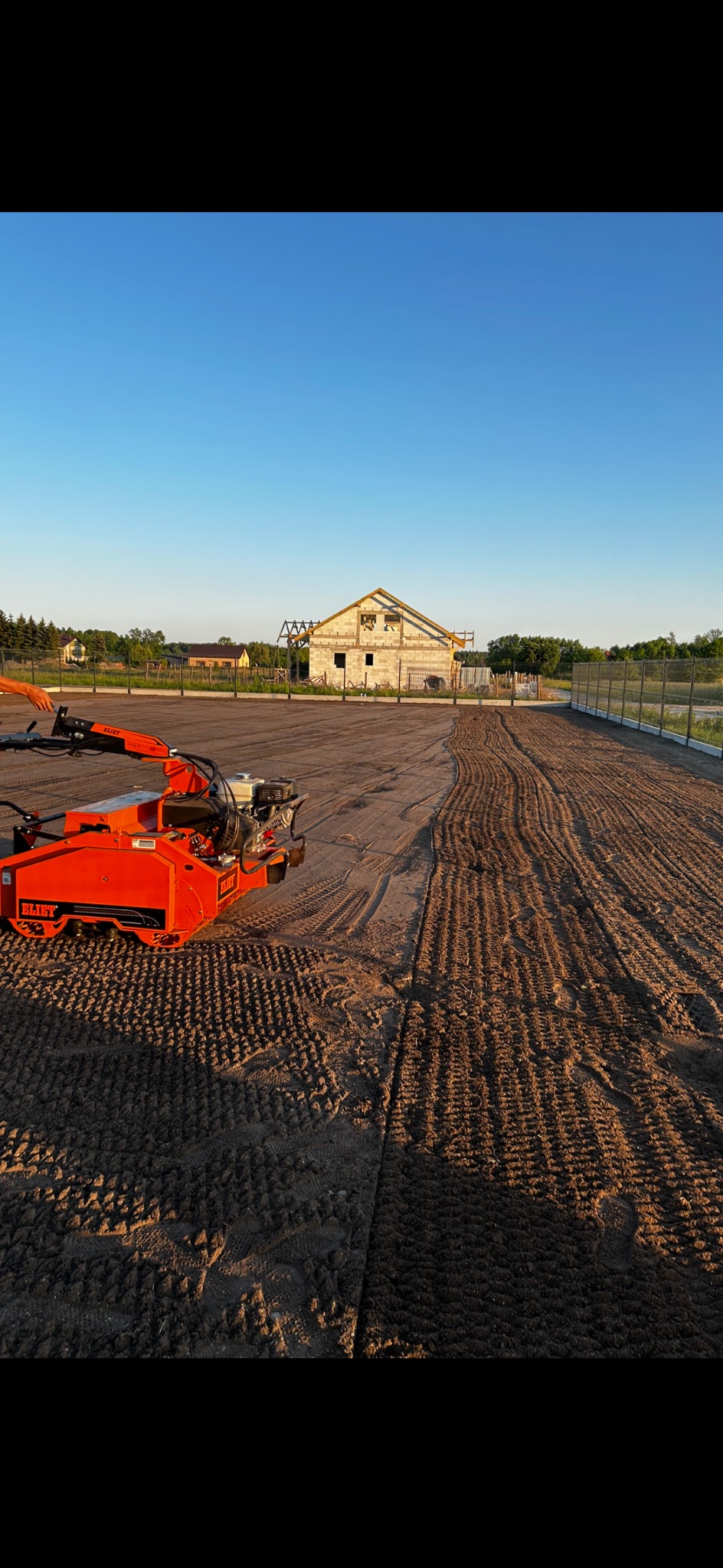 Pomarańczowa maszyna marki ELIET wyrównuje teren pod budowę, widoczne ślady opon i wzór na ziemi, w tle budynek w trakcie budowy i błękitne niebo.